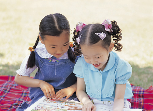Two girls reading a book together outdoors on a blanket 